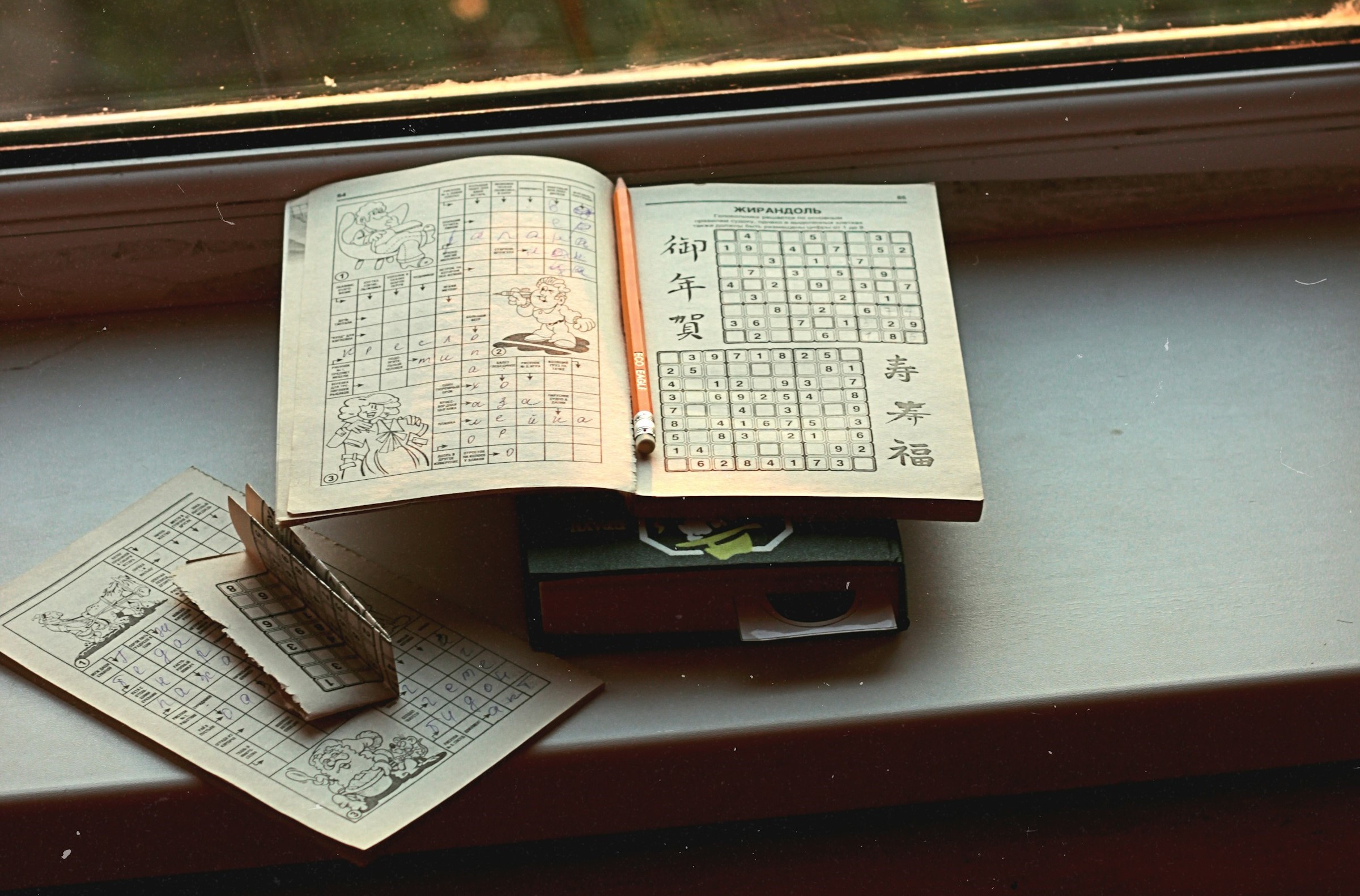 A stack of books on a window sill. On top is a puzzle book featuring Sudoku puzzles, with a pencil.