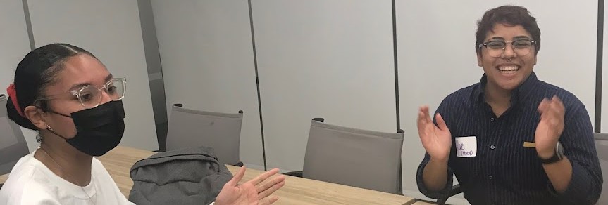 Two students wearing glasses across from each other at a table, one on the left with a mask on and hand up ready to clap, on the right one unmasked smiling clapping