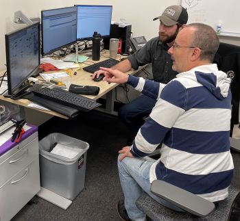 Two men sit together looking at computer monitors