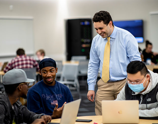 An instructor in a blue button up and yellow tie stands and speaks with three students seated around a table.