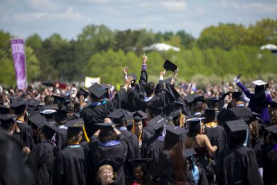 ​ A crowd shot of UAlbany graduates in black graduate robes and mortar board caps celebrating Commencement with their arms raised in the air.