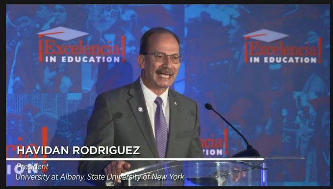 A screenshot of President Rodríguez standing alone at a podium in a gray suit and addressing the audience at the Seal of Excelencia announcement. 