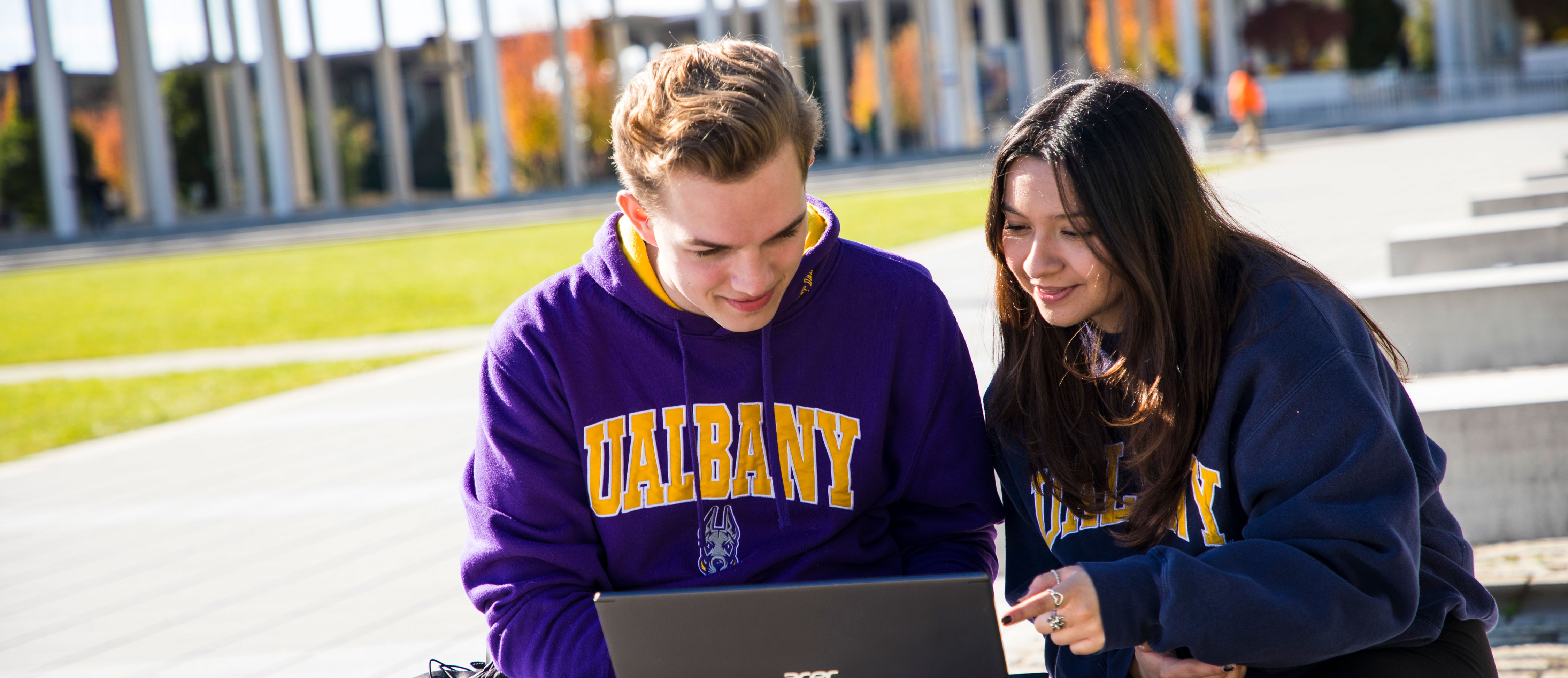 Two students wearing UAlbany sweatshirts sit on a bench outdoors on the Uptown Campus and look at a laptop together.