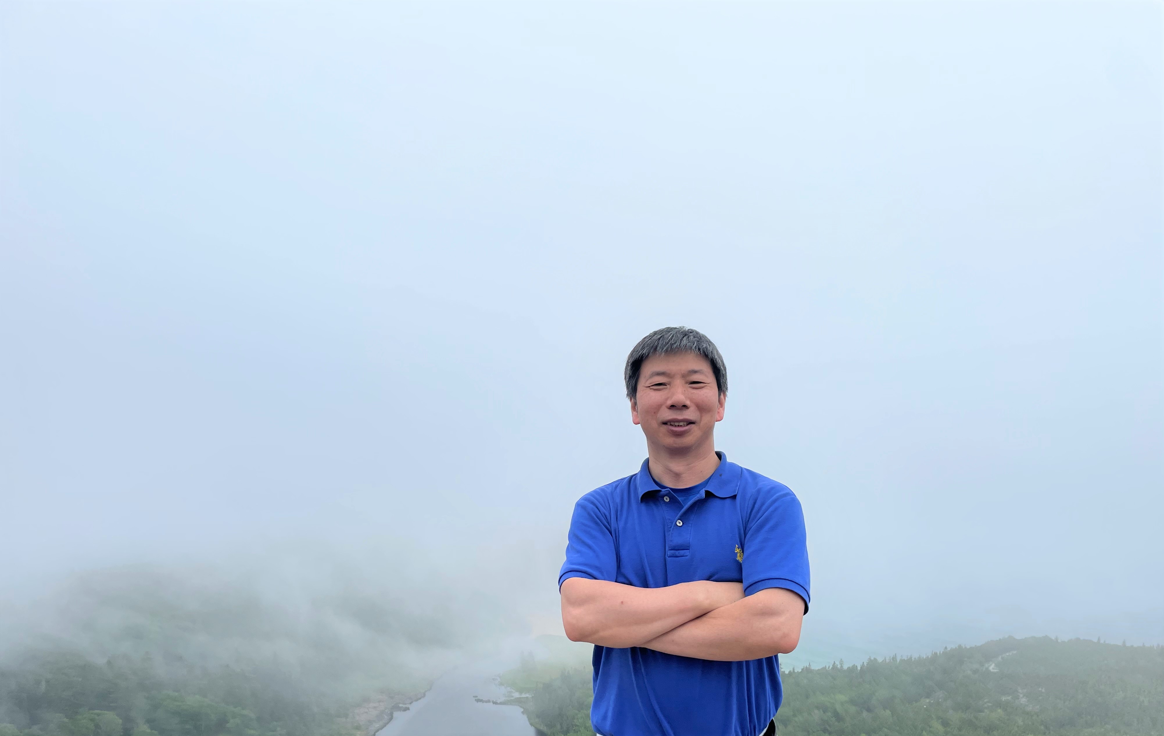 ASRC's Fangqun Yu stands in front of a foggy road in a blue polo with his arms crossed.