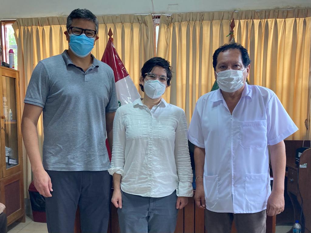 Beth Feingold stands between two colleagues in an office in Peru. The three are standing in front of a curtained window; the curtain is cream-colored and a portion of the national flag of Peru is visible behind them. 