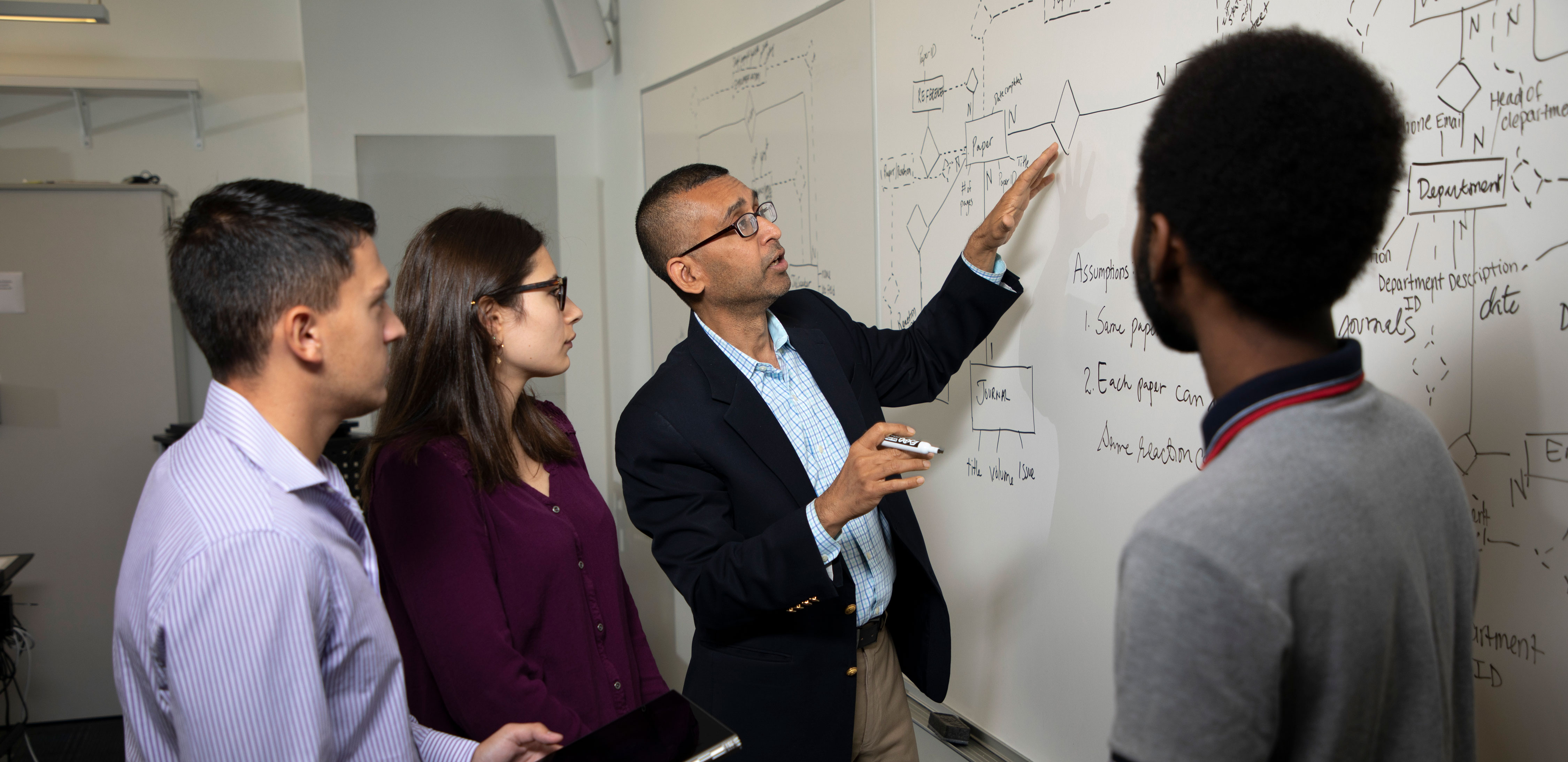 An instructor standing with three students gestures toward and writes on a white board.