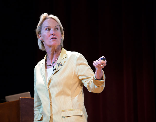 Frances Arnold stands onstage wearing a yellow blazer and holding a powerpoint pointer in her left hand.