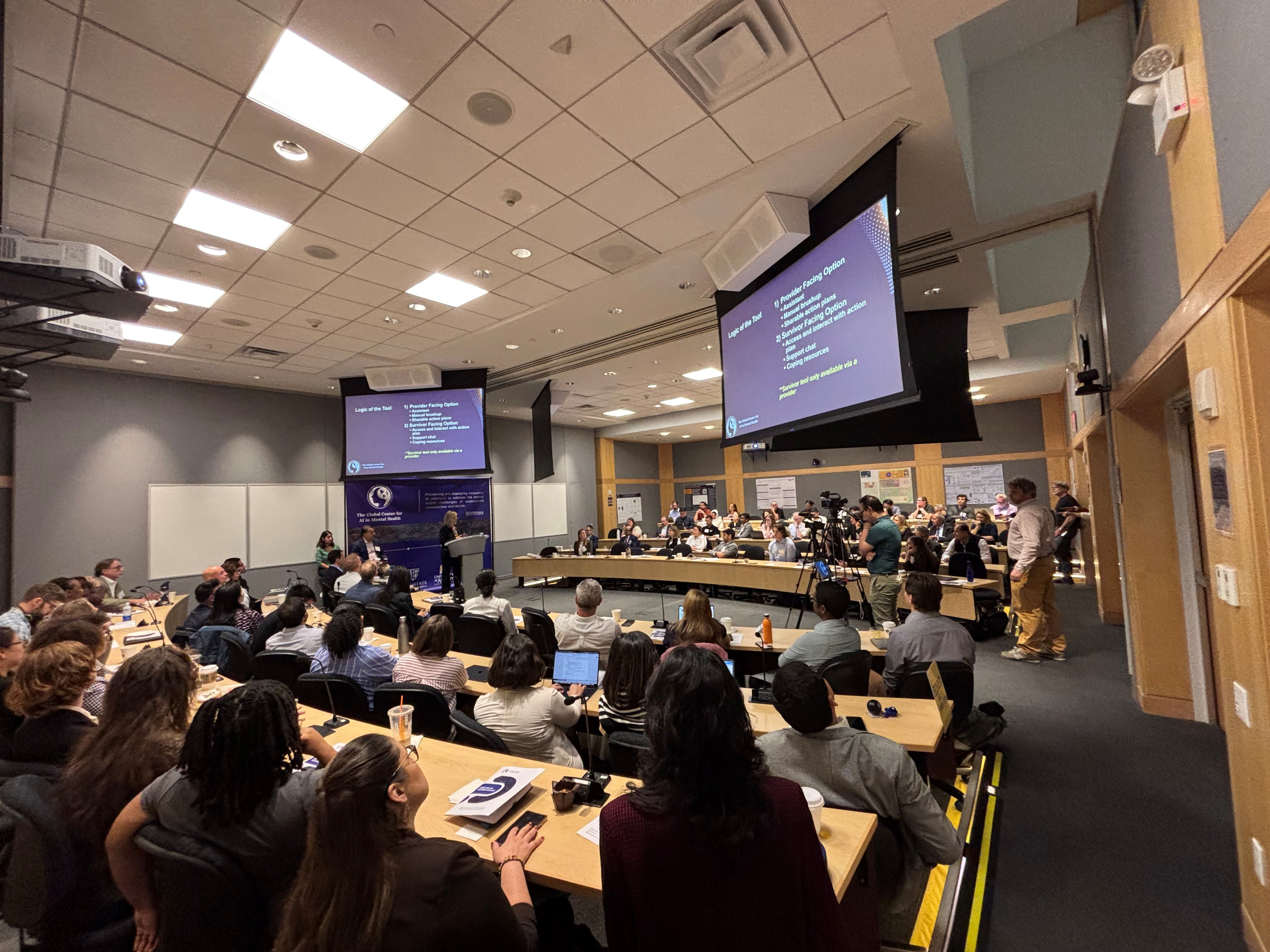 Over one hundred people convene in a brightly lit conference room. All are seated at long wooden desks arranged in rows. Two large projector screens hang from the ceiling.
