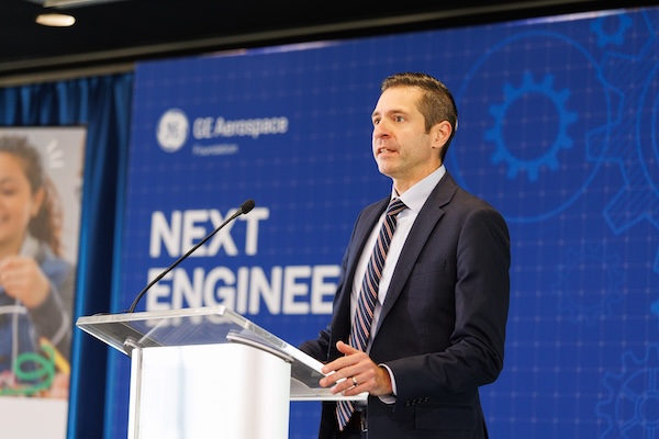 A man in a suit stands at a podium in front of a blue screen that reads "Next Engineers"