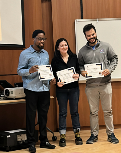 Three PhD candidates smile holding certificates in the D’Ambra auditorium in the RNA Institute. From left to right: Emmanuel Edem Adade, Michelle Urman, and Jesus Frias. A whiteboard and projector are in the background. The walls are covered in wood paneling.