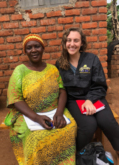 Giana sits outside with a woman in Uganda. Behind them is a brick wall. 