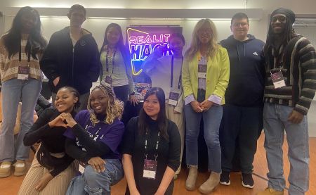 A group of 10 people stand around a neon sign reading Reality Hack