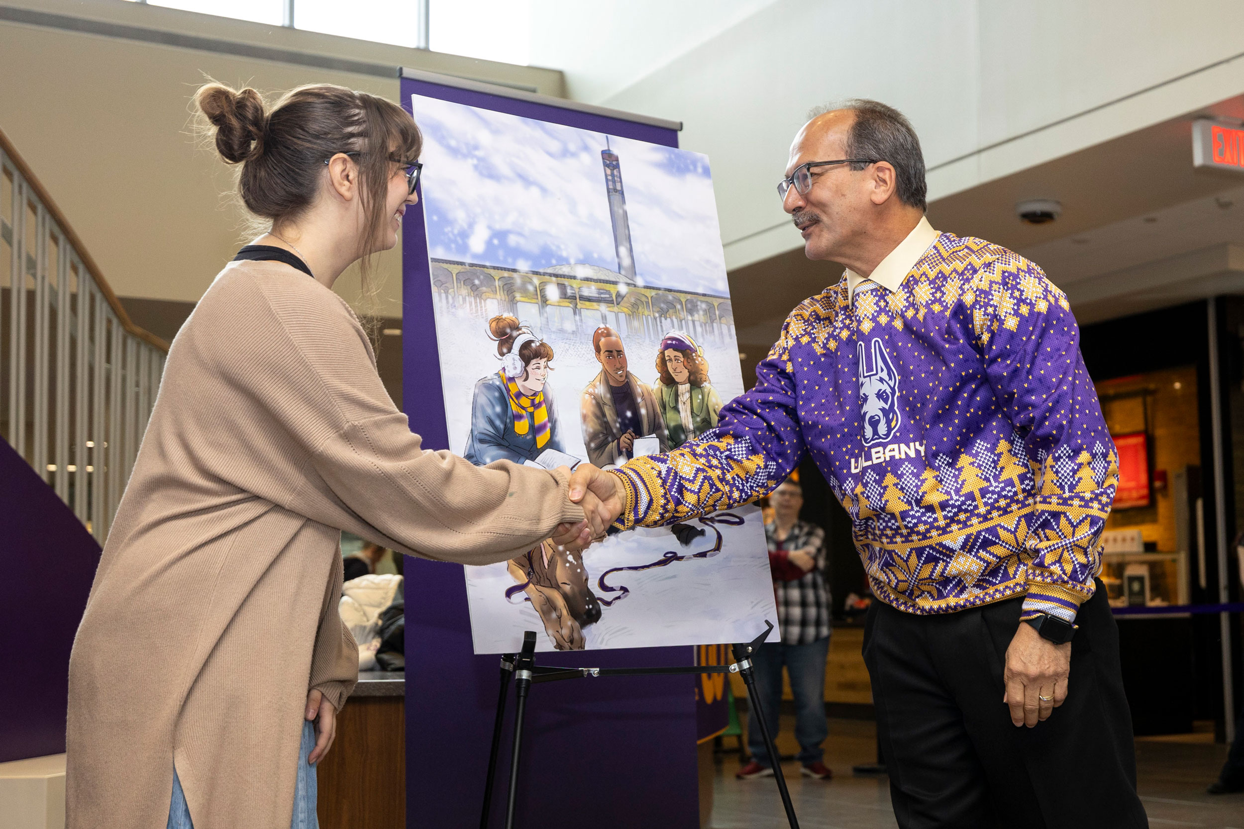 UAlbany President Havidán Rodríguez shakes a student's hand as they stand in front of an easel holding the winning 2024 Holiday Card Art Contest.