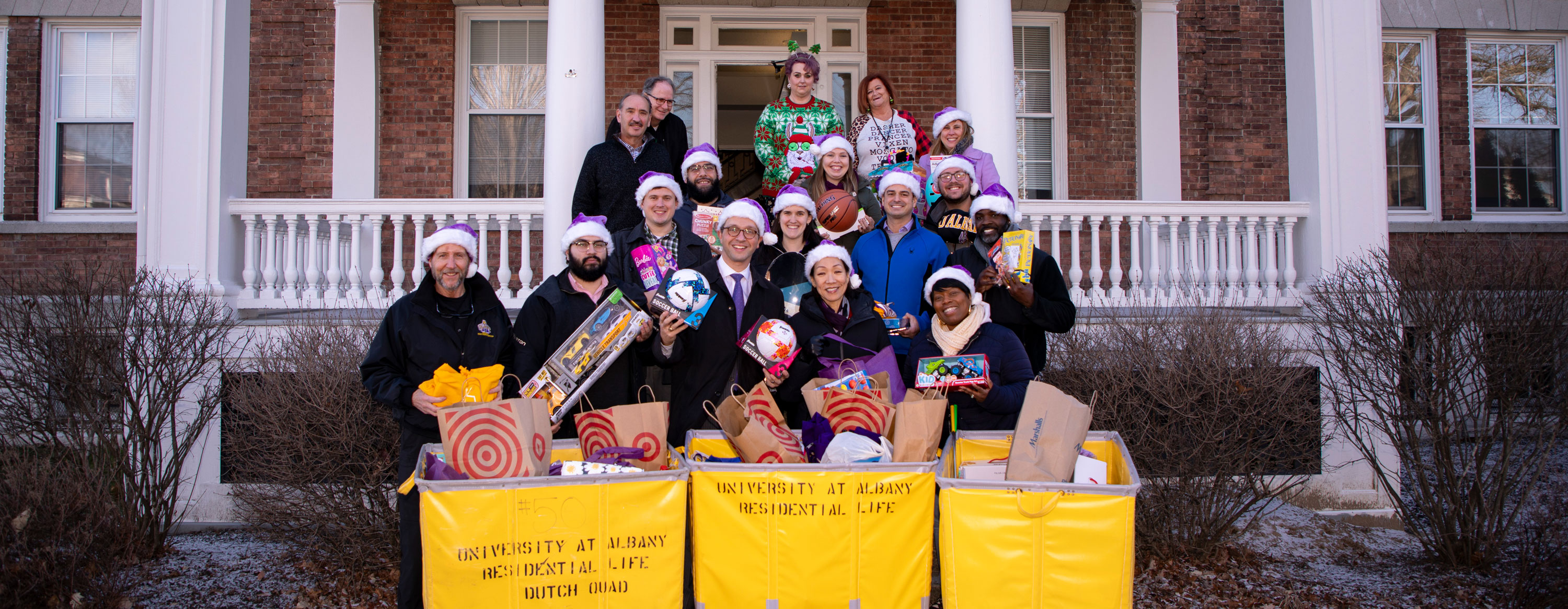 Seventeen UAlbany employees wearing purple Santa hats smile and pose with three carts full of holiday gifts.