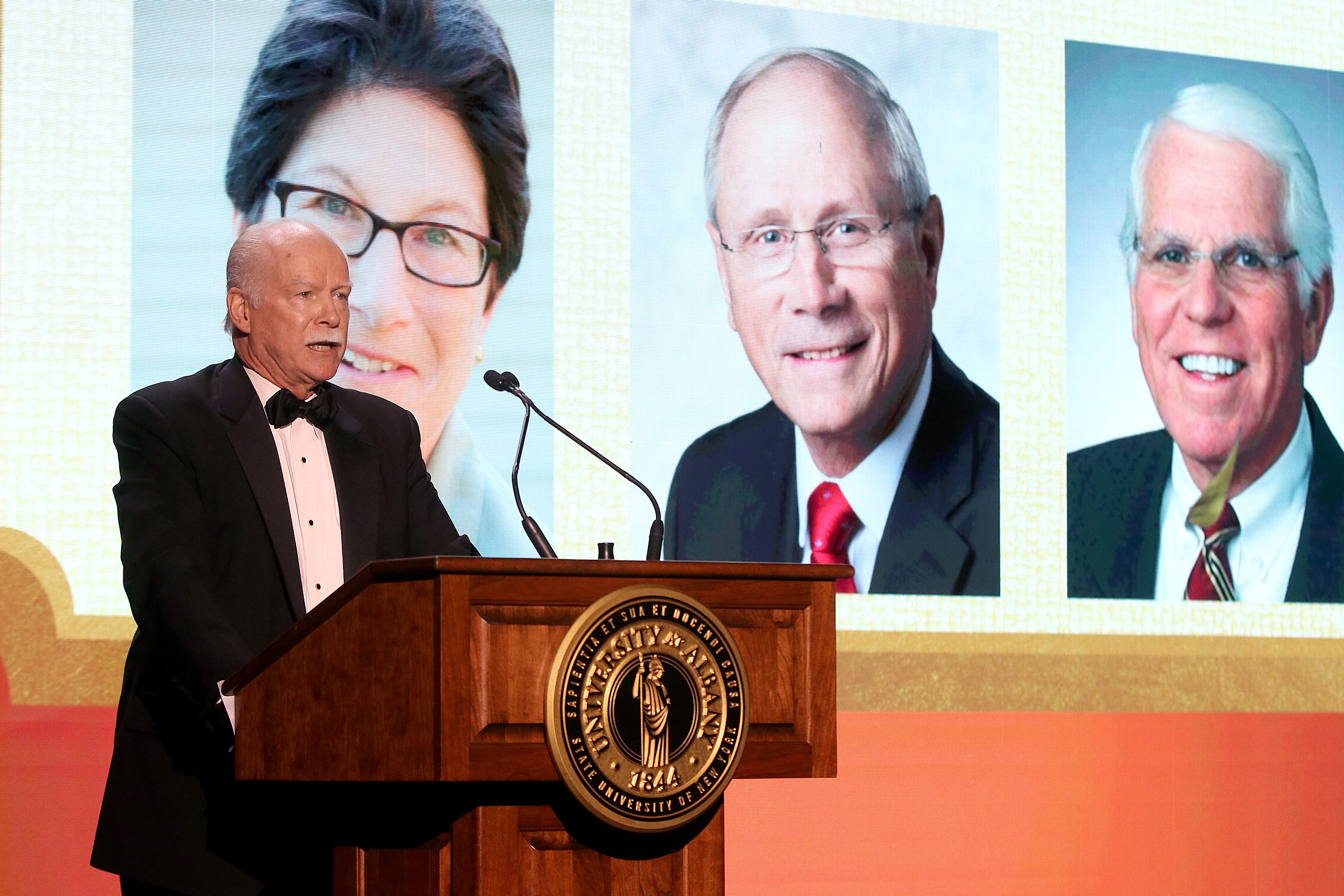 A man in a tuxedo speaks at a lectern framed in the background by the images of three faces projected on a screen behind him.