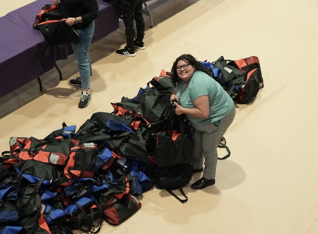 A woman with dark curly hair and glasses stands beside a large pile of blue and red duffle bags.