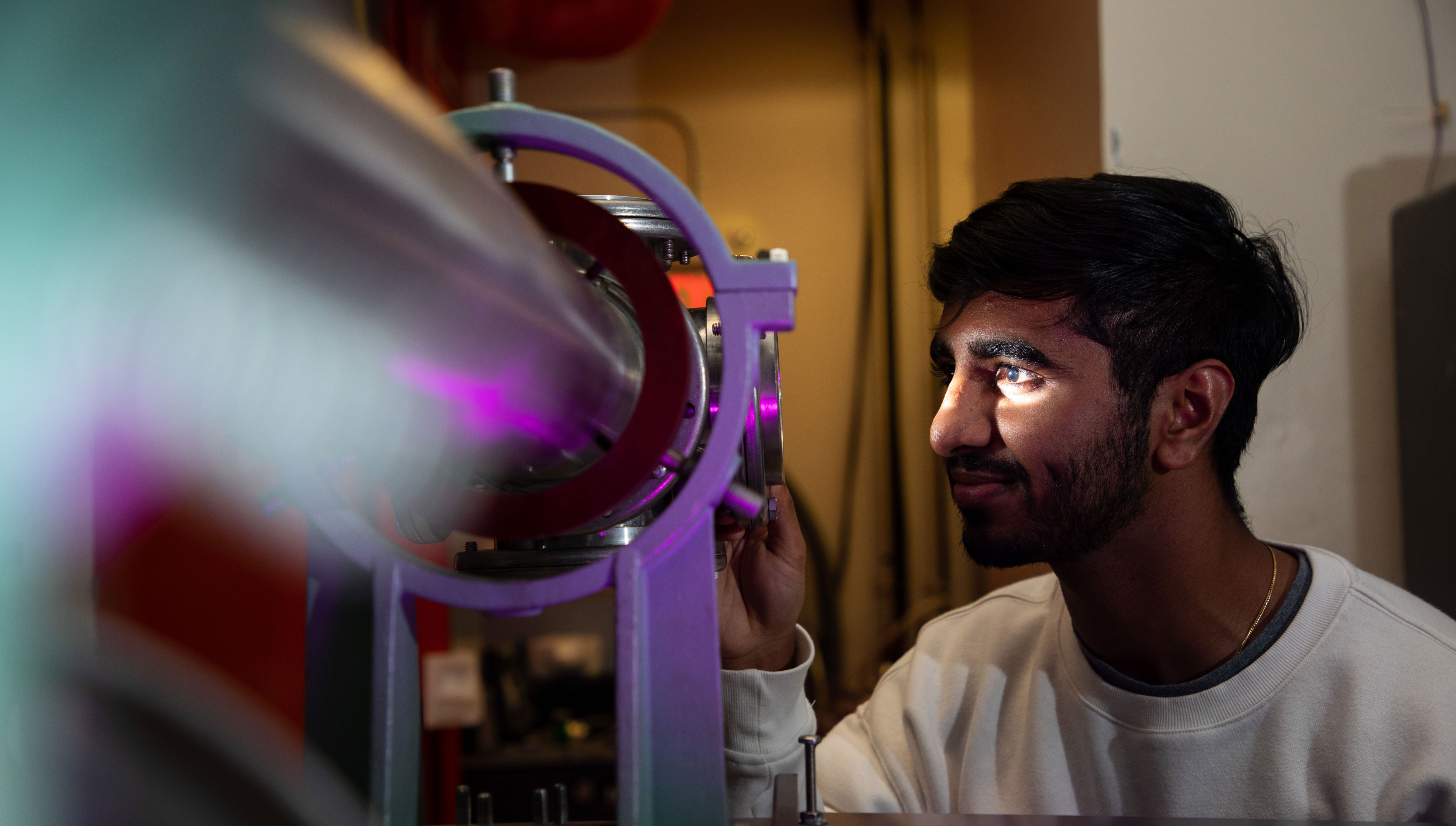 A researcher smiles as he looks into a piece of equipment lit up in purple inside the Ion Beam Lab.