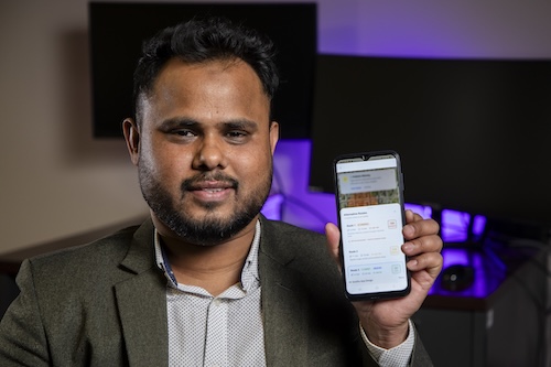 A smiling man in a gray suit jacket holds up a phone to demonstrate an app