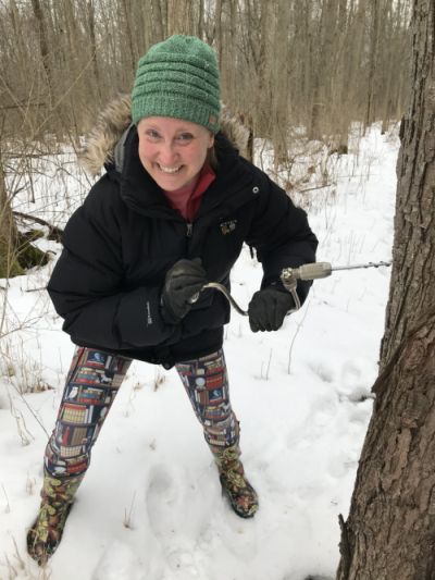 Jessica Castner is dressed in warm winter clothes, drilling a maple tap into a tree in a snowy forest. 