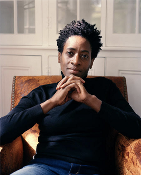 A seated woman with short black hair wears a black turtleneck and stares into the camera, her hands crossed beneath her chin.