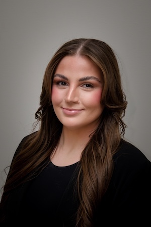 A woman with long brown hair in a black shirt smiles for a portrait against a gray backdrop.