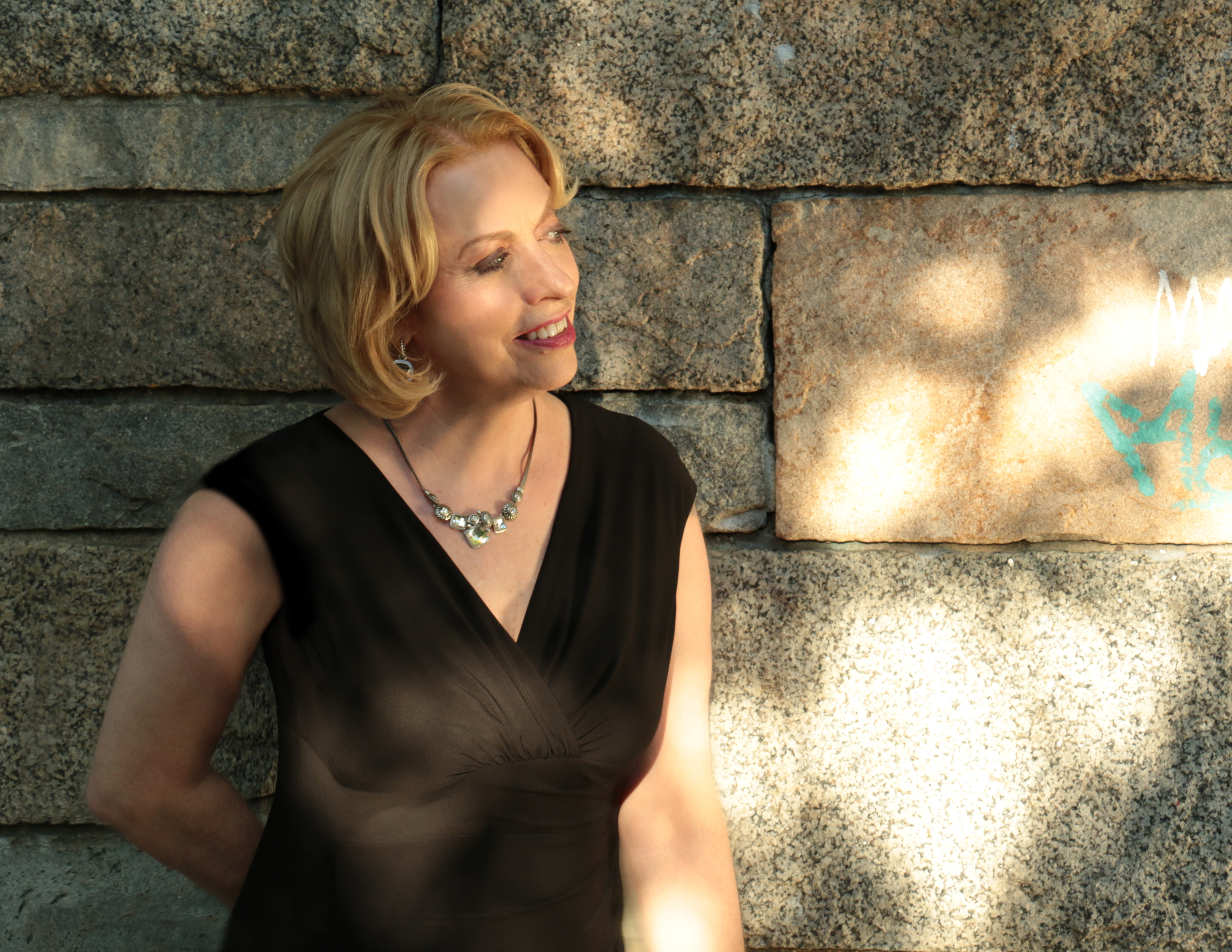 woman with short blonde hair in black dress and silver necklace smiles and leans against a stone wall dappled in sunlight