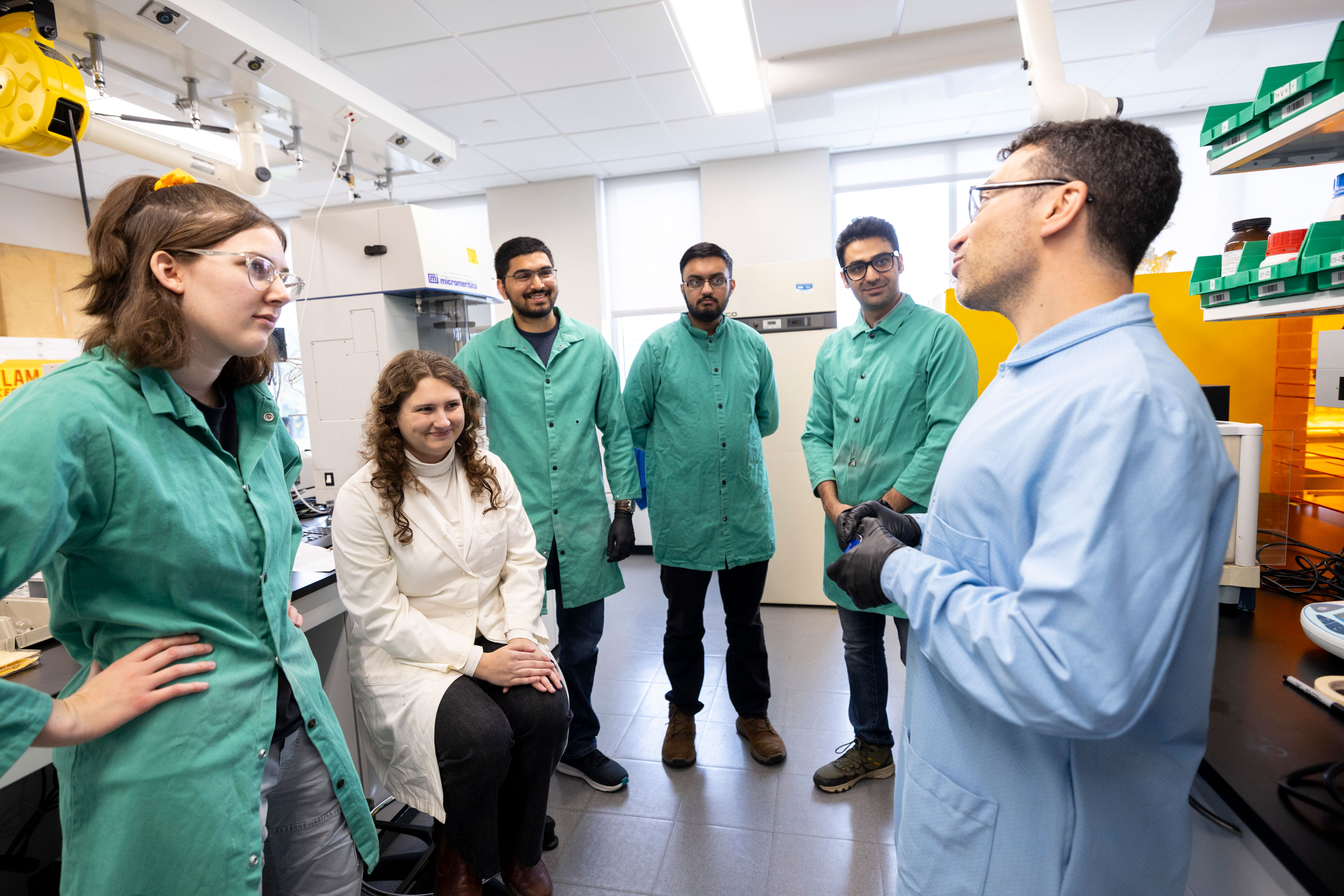 A chemistry researcher in a blue lab coat speaks to five members of his lab team, four of whom wear green lab coats and one of whom wears a white lab coat.