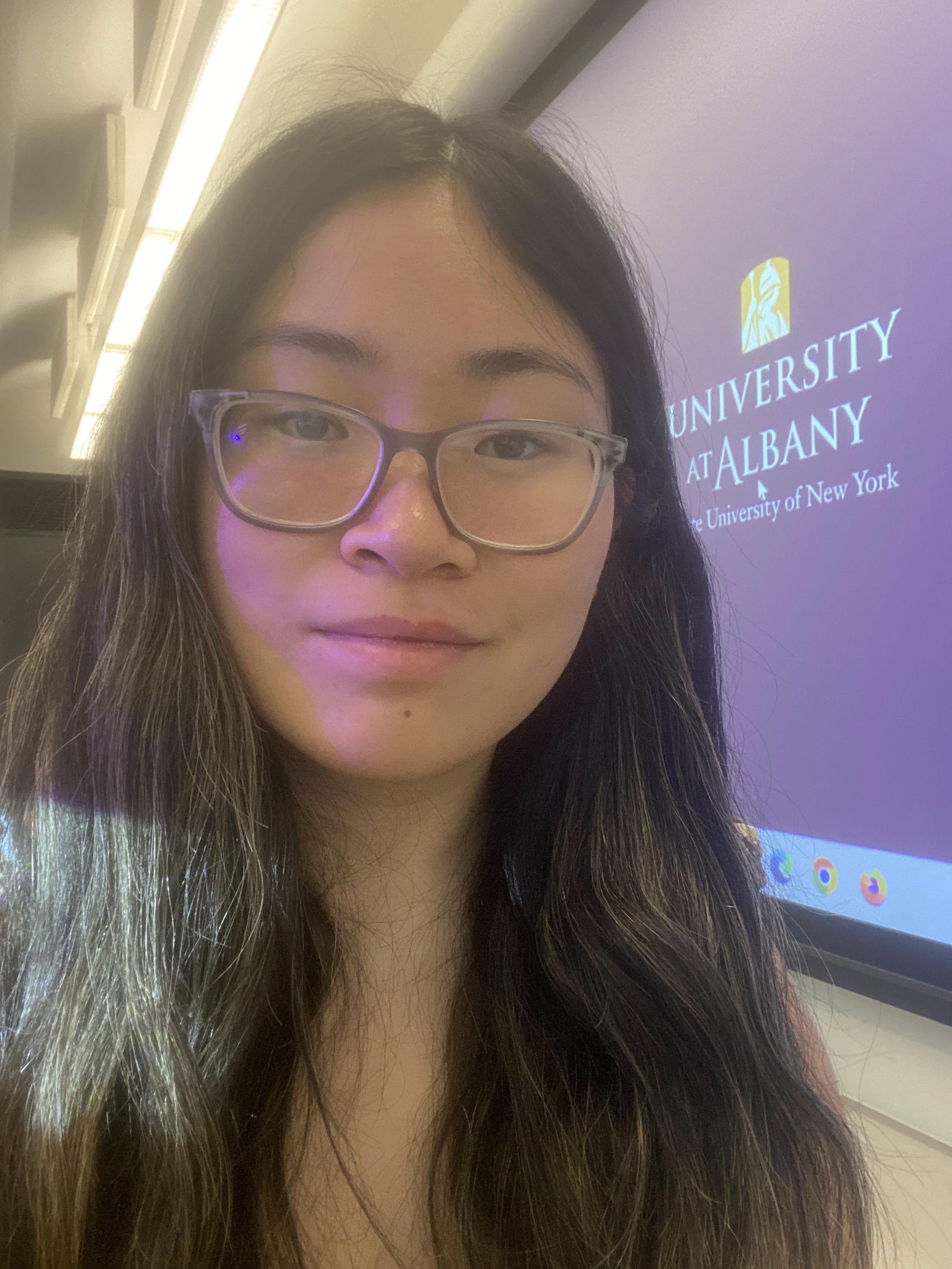 A woman with long dark hair and glasses smiles in a classroom with a purple screen reading "University at Albany State University of New York" in the background