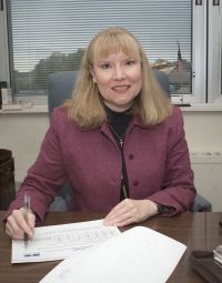 A woman seated at a desk smiles for the camera, pen poised in hand above some papers.