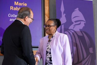 A man in a black suit shakes the hand of a woman in a pale purple jacket in front of a poster of the goddess Minerva