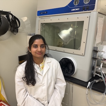 A woman with long dark hair in a white lab coat sits next to lab machinery