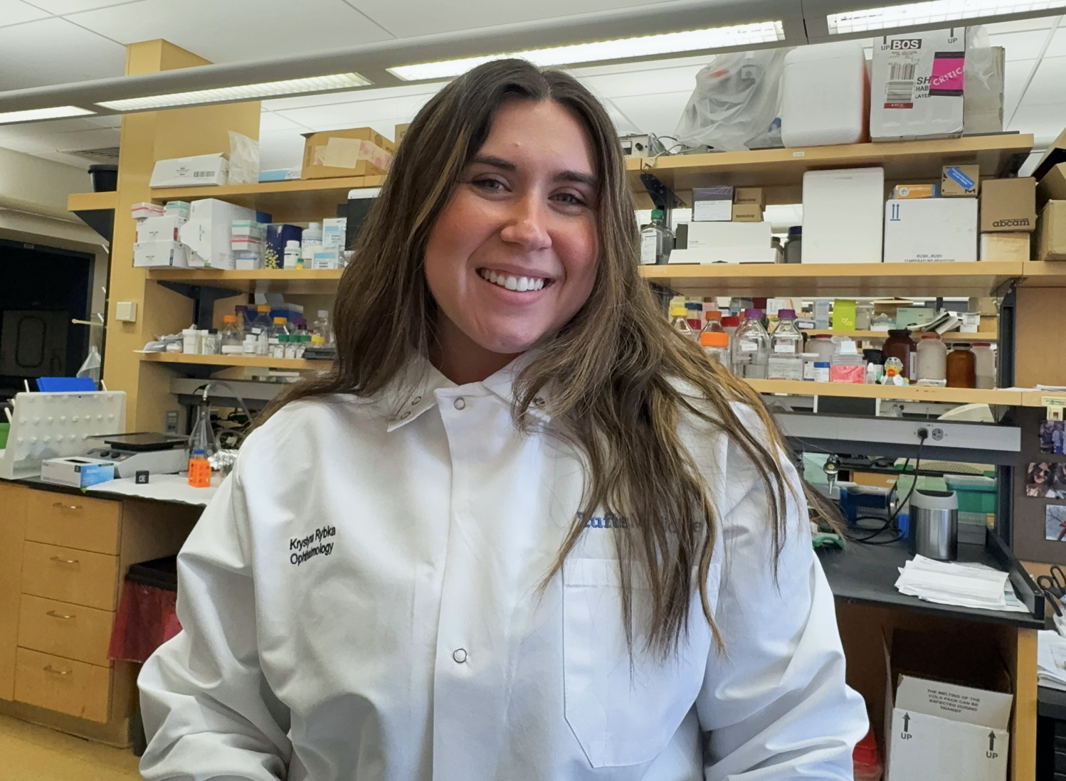 a smiling woman in a white lab coat stands in a research lab