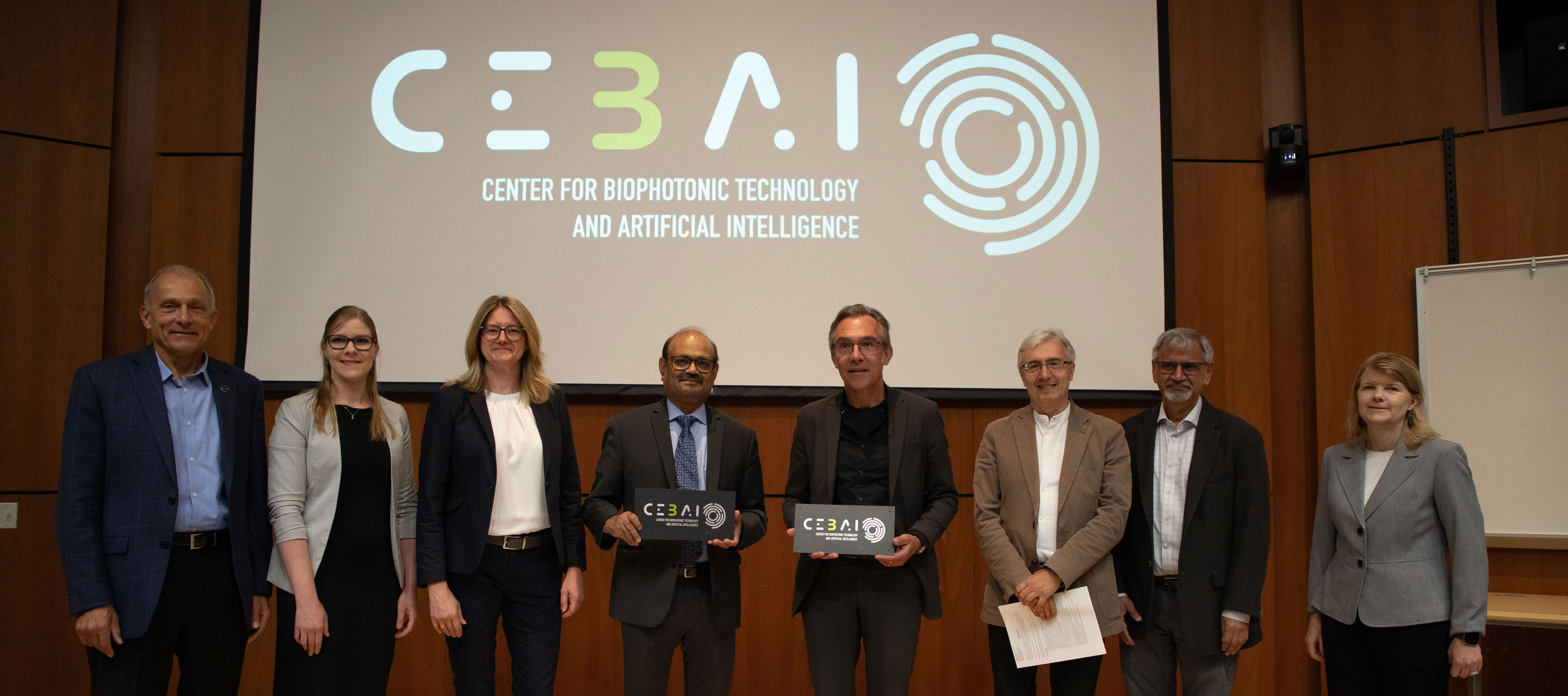 Eight officials pose for a photo in front of the Center for Biophotonic Technology and Artificial Intelligence (CeBAI) logo at the center's launch event.