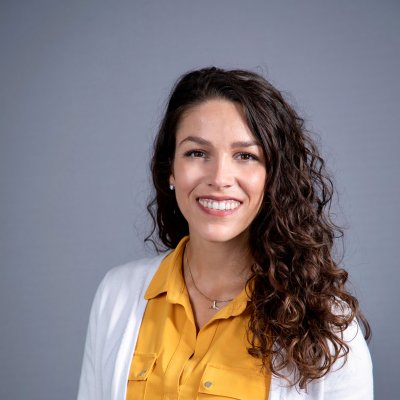 A woman with long curly brown hair wearing a white cardigan over a yellow blouse smiles for a portrait against a gray backdrop