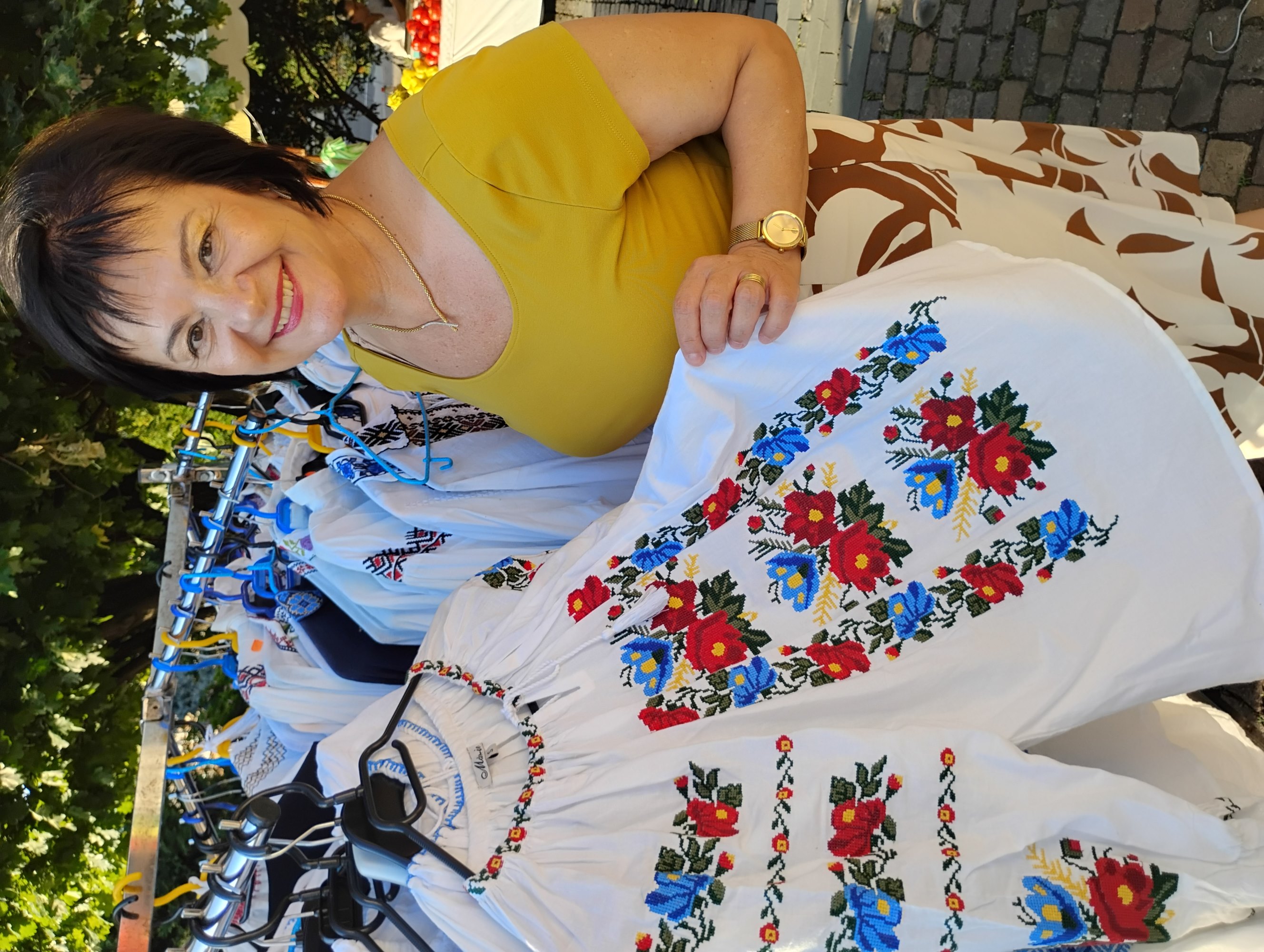 A woman in a yellow blouse holds a shirt in a traditional floral pattern from Transylvania, Romania, outside in an open-air shop in Cluj-Napoca, Romania.