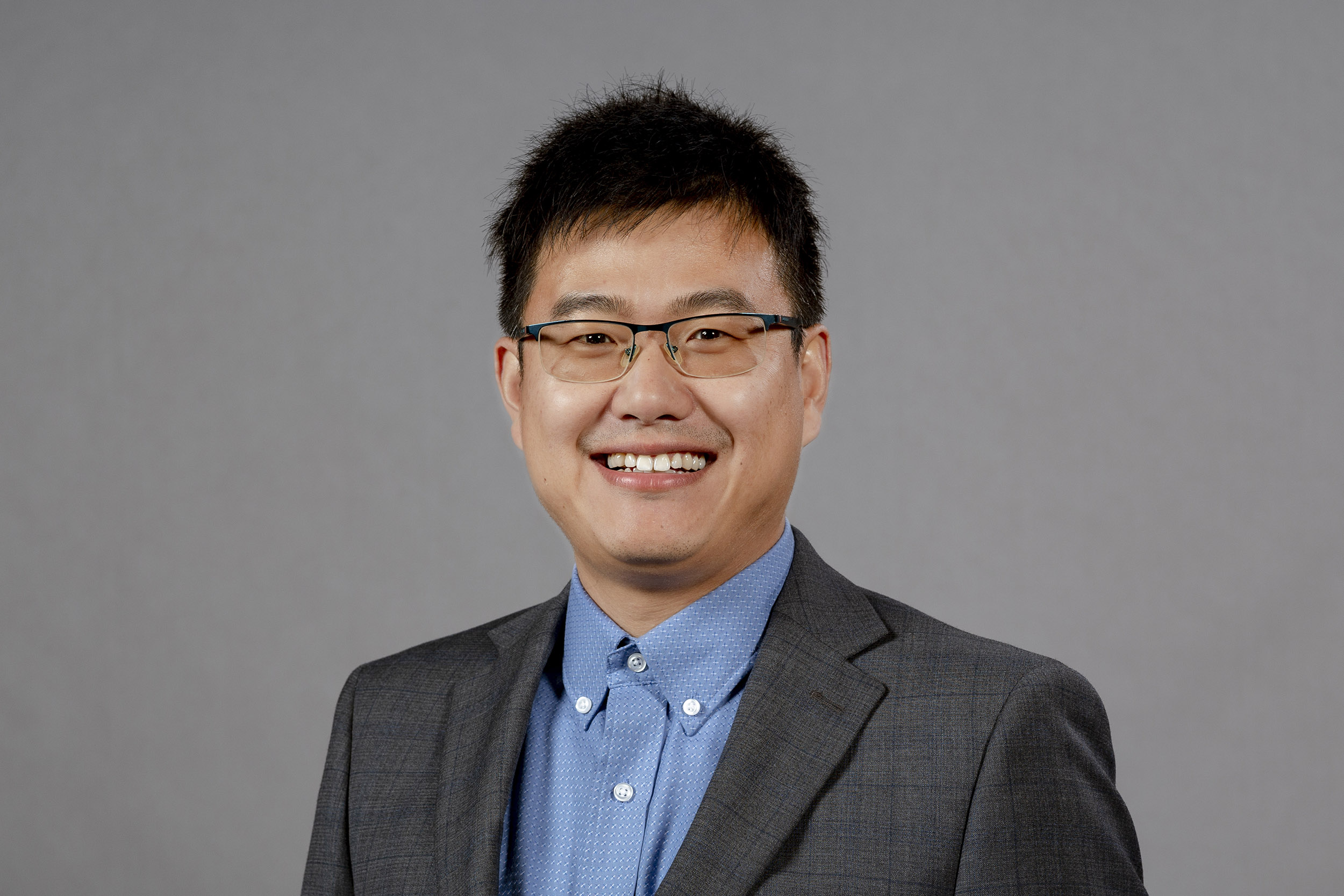 A portrait of a man with dark hair, glasses, a grey suit and a blue buttoned down shirt.