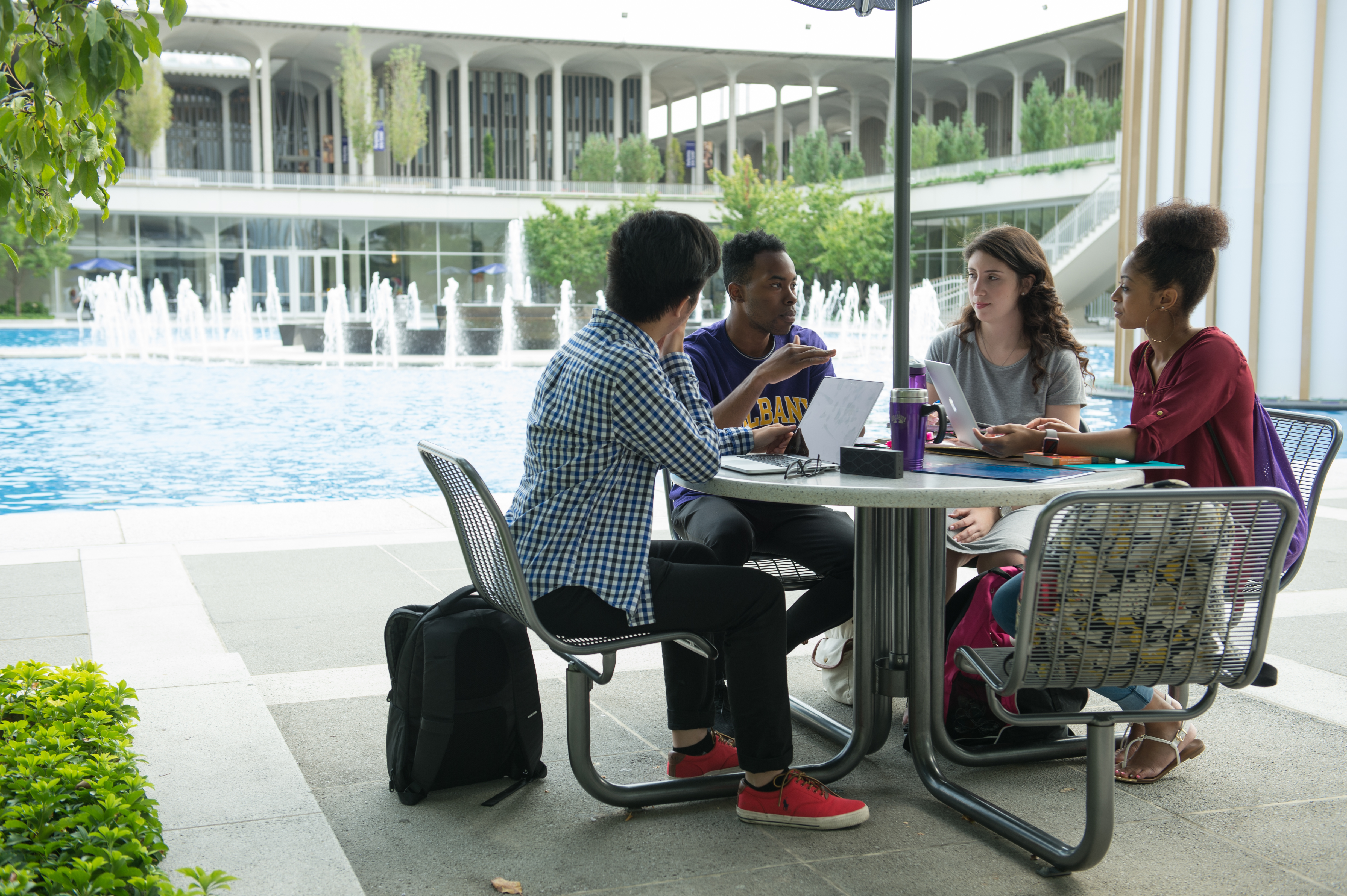 Two male and two female students sit talking at a table with the campus main fountain in the background talking 