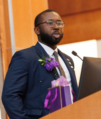 A man in a blue jacket with purple floers on the lapel speaks at a lectern