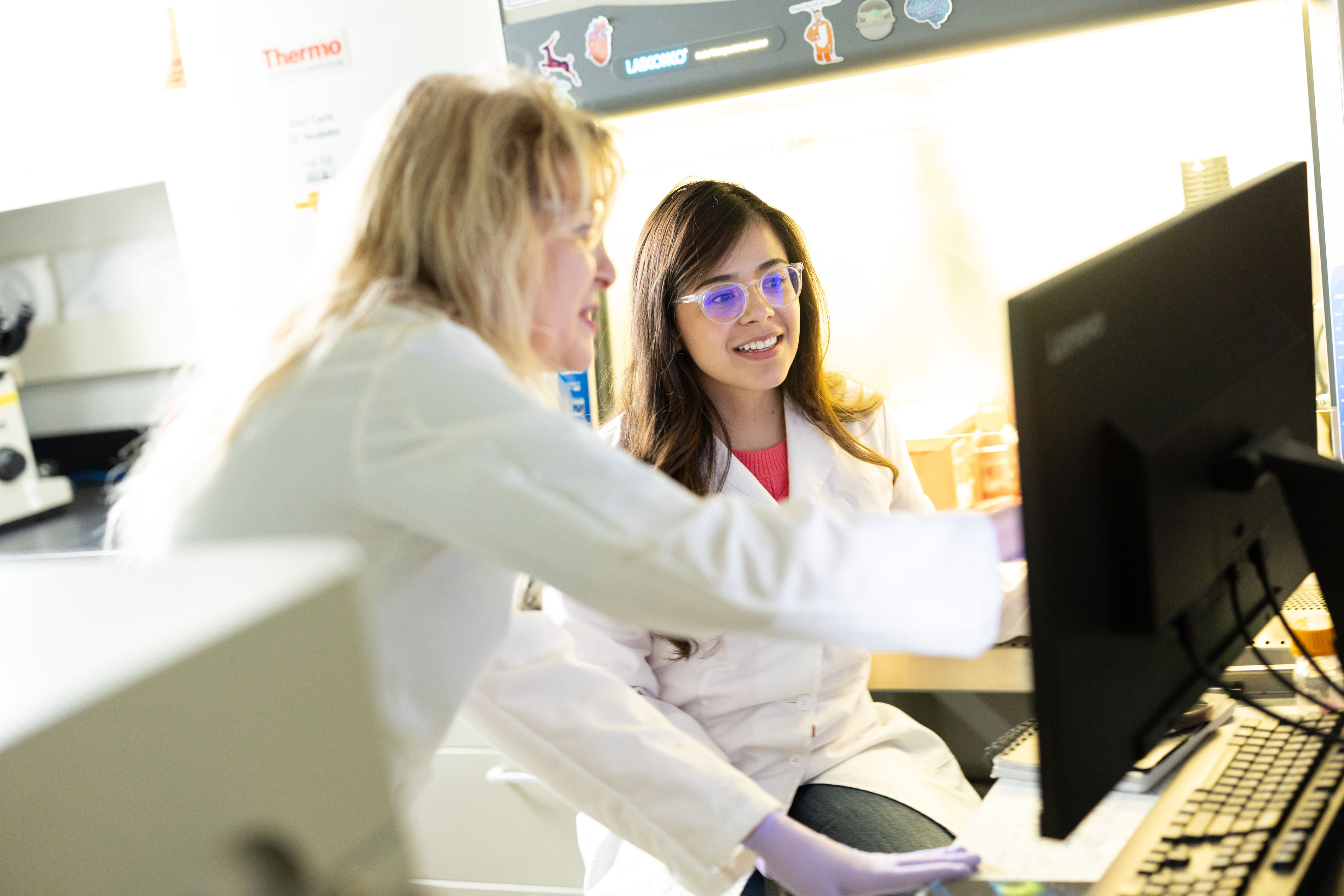 A researcher and a lab member wearing purple gloves and white lab coats sit inside a lab looking at a computer screen together.