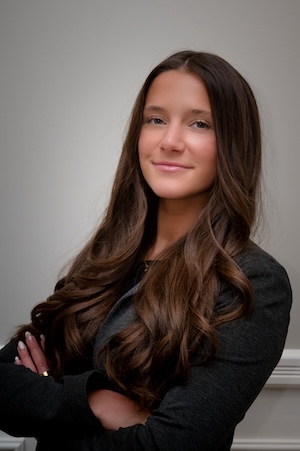 A young woman with long brown hair in a black blazer folds her arms and smiles for a portrait against a gray backdrop.