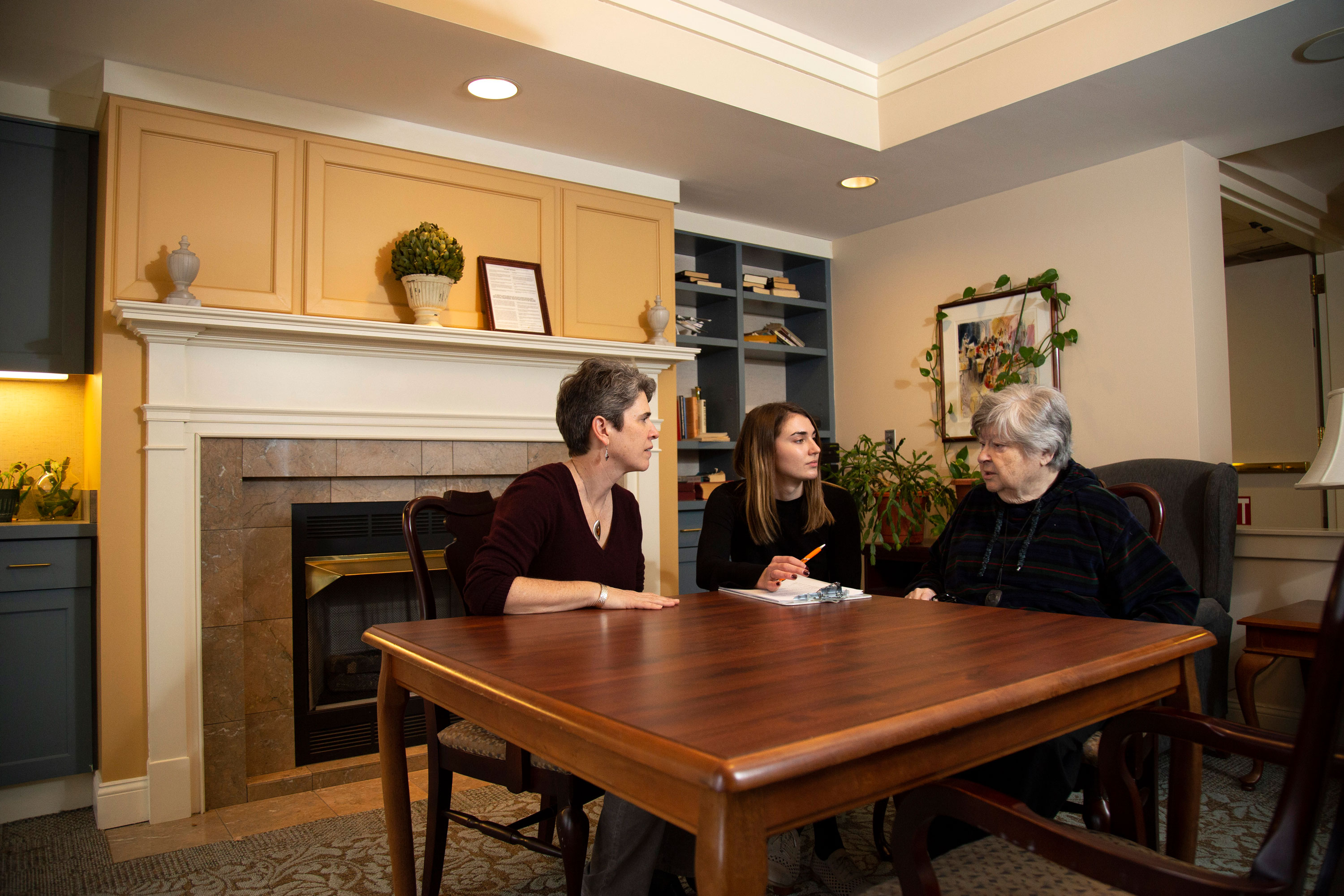 Three adults of varying age sit at a table inside a sitting room and speak.