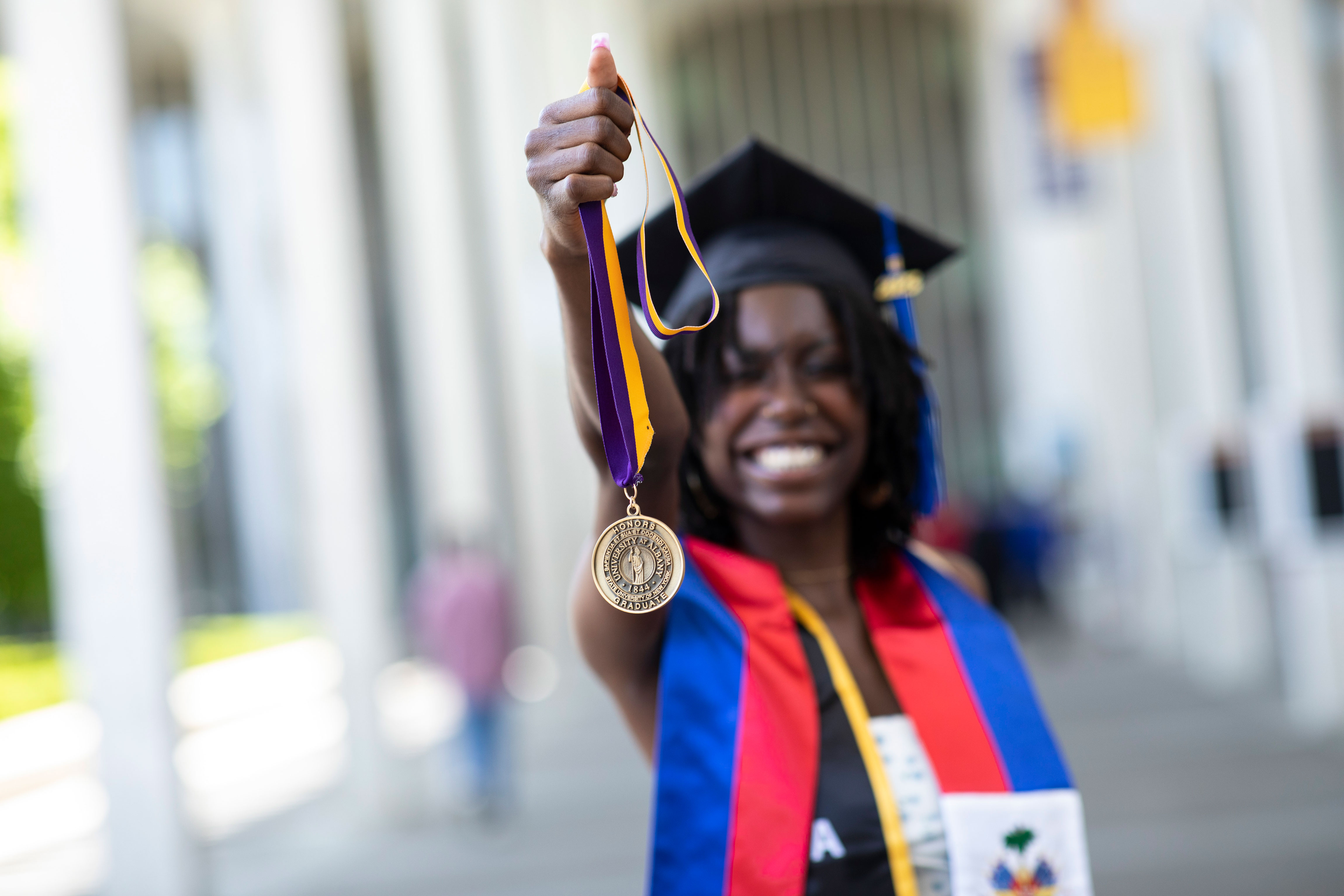A student wearing a graduation mortar board smiles and holds up an honors medallion.