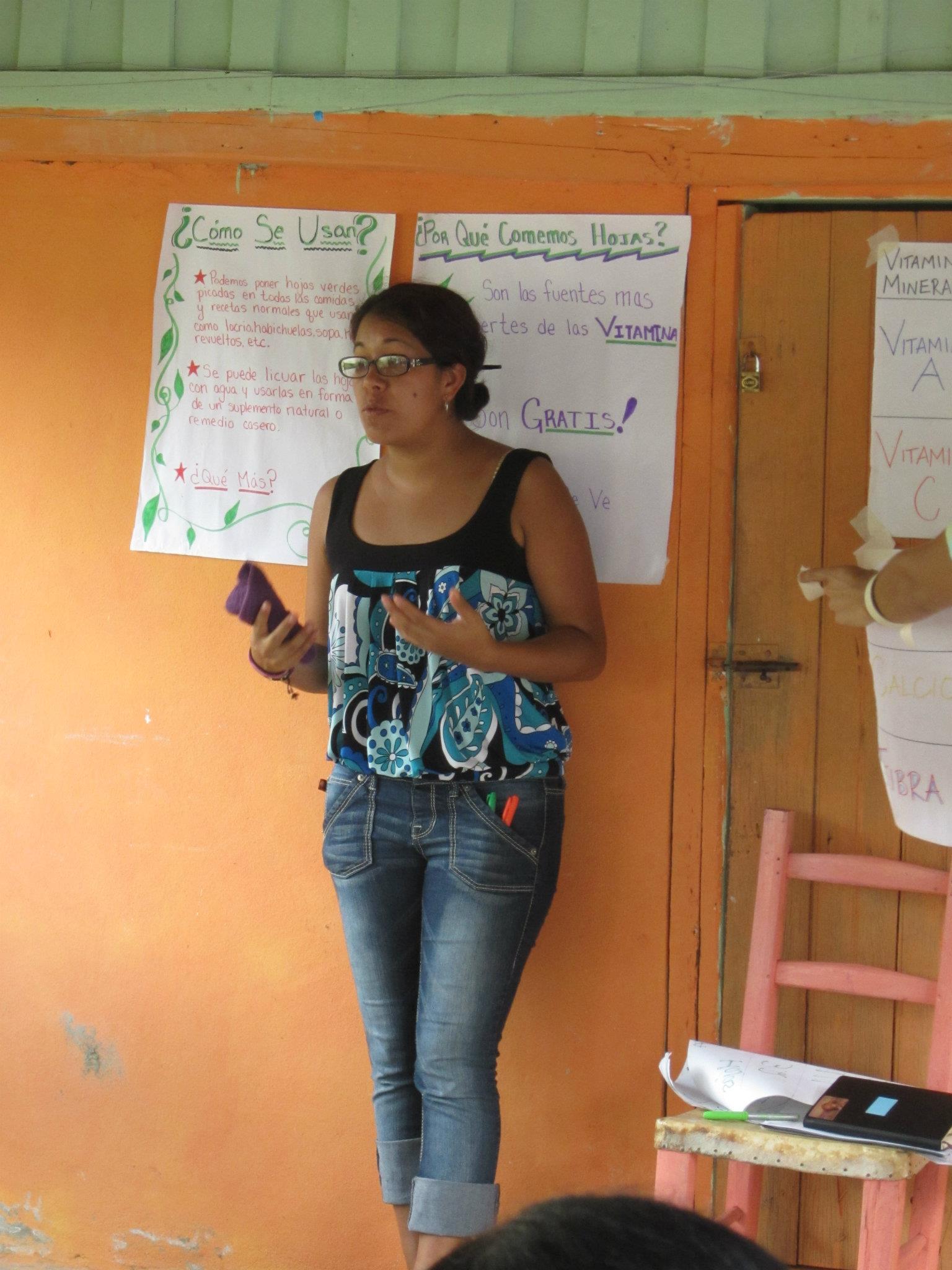Melissa stands in front of an orange wall, teaching public health.