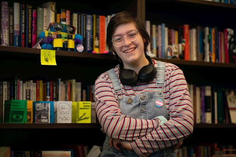 Melody Palmer in overalls and a striped shirt, stands in front of a book case