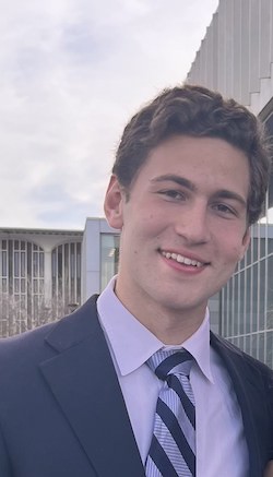 A young man with short light brown hair smiles in a suit jacket and tie on the UAlbany campus.