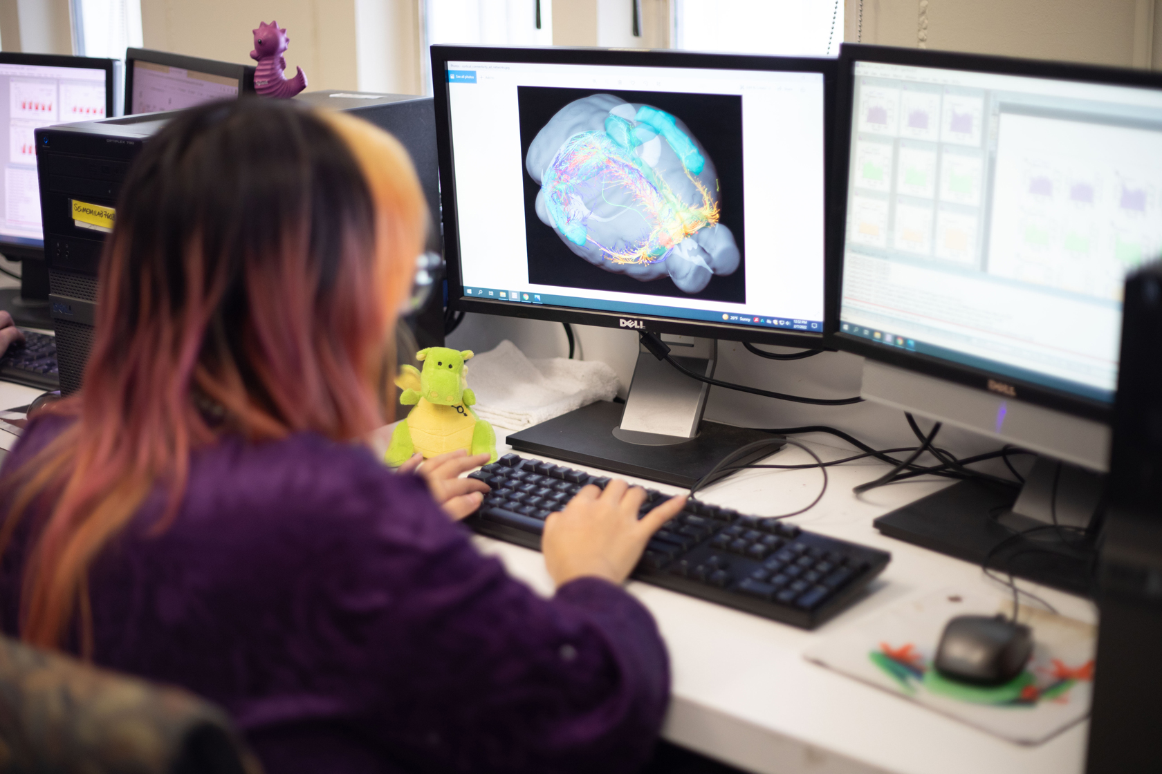 A student in a blue lab coat and googles completes research at a workstation, surrounding by shelving and scientific equipment