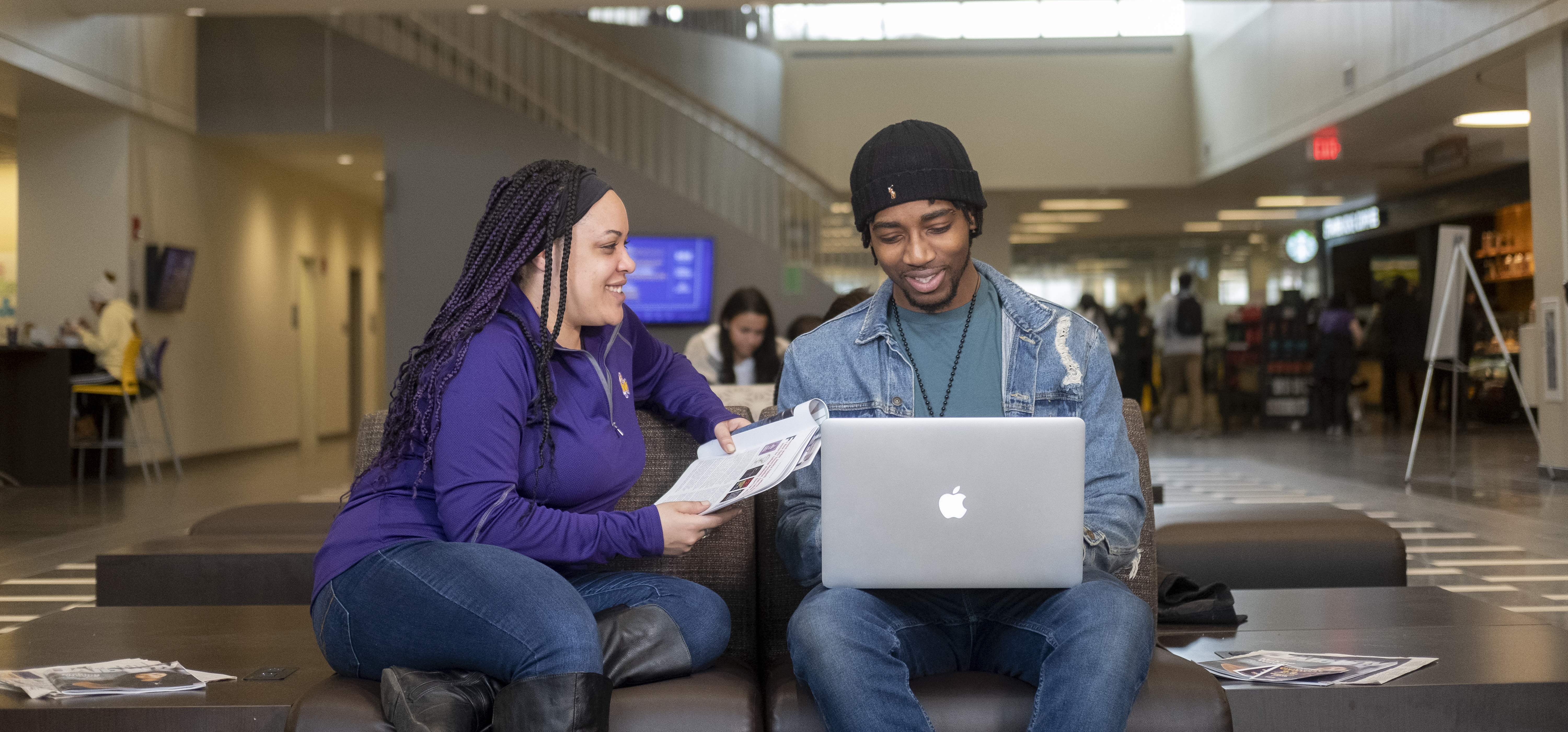 Two smiling students sit in Campus Center holding a laptop and some papers.