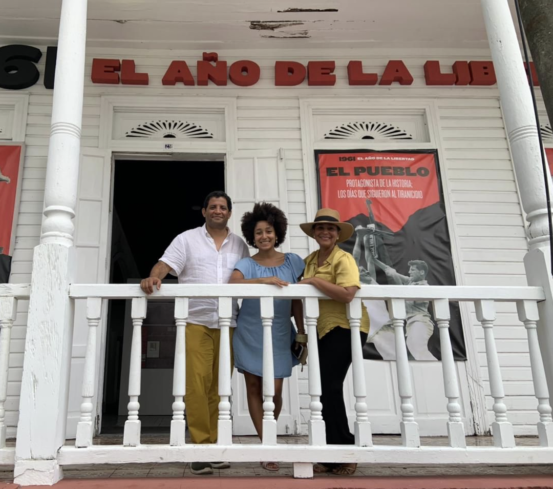 Three people lean against a balcony railing and pose for a picture in front of posters highlighting an art exhibition called "1961: El año de la libertad"