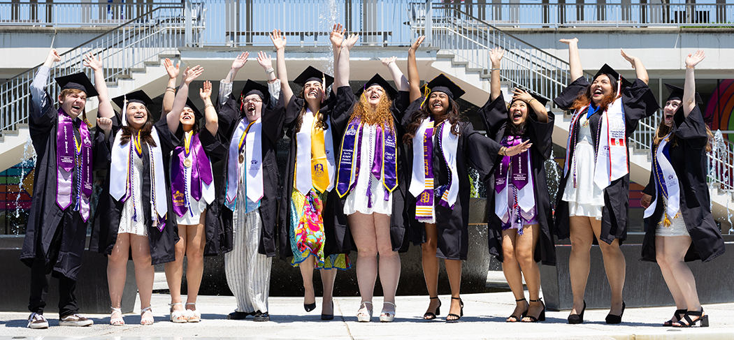 A large group of UAlbany graduates cheers happily, their arms raised.