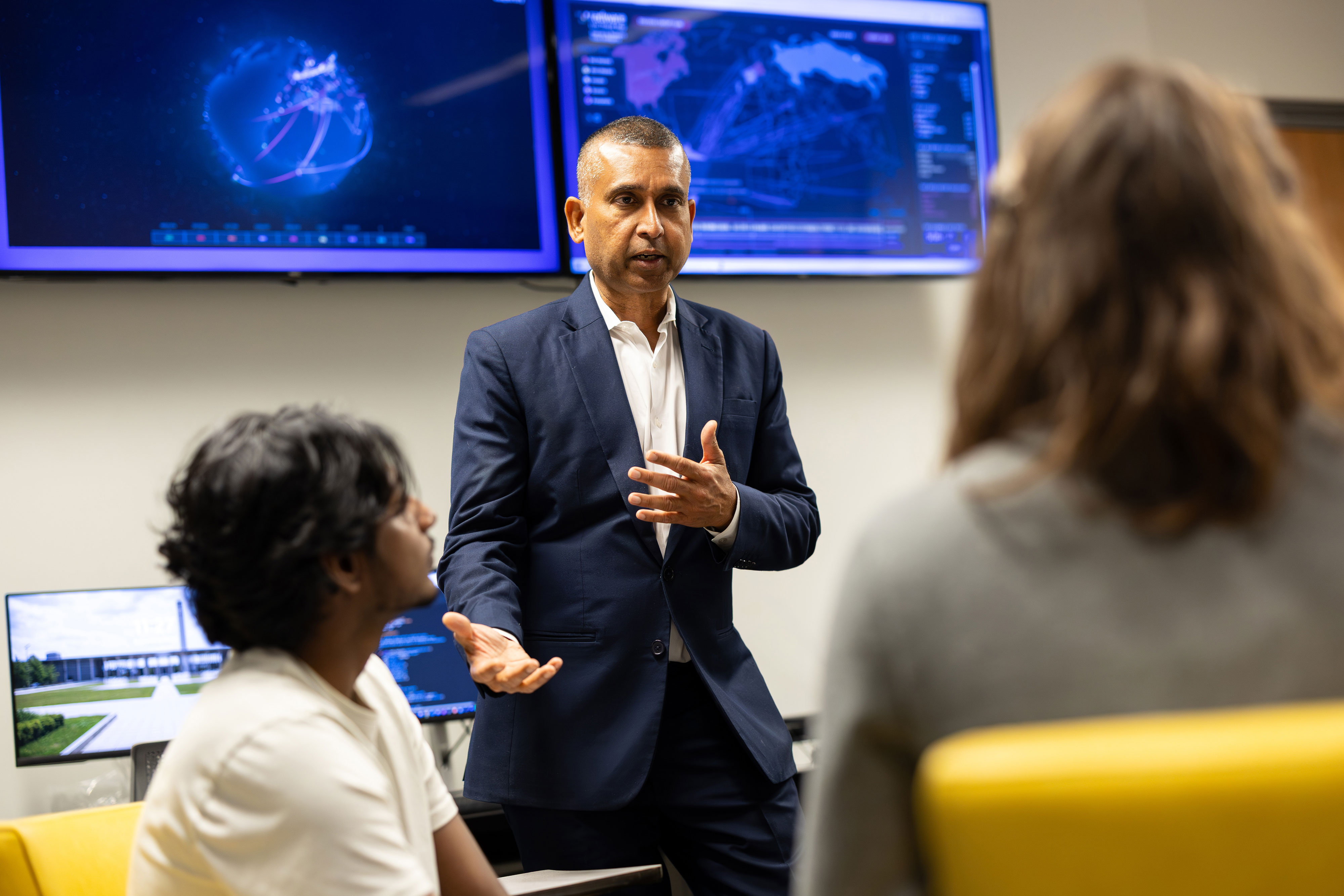 A researcher standing in front of several display screens gestures as he speaks to a group of people.